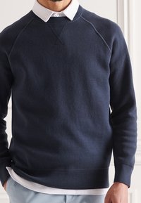 Felpa blu navy realizzata in tessuto a maglia, con scollatura a girocollo, polsini e orlo a coste, indossata sopra una camicia bianca con colletto.