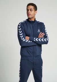 Navy trainingspak van gladde stof, met witte chevronstrepen langs de armen en benen. Inclusief een zipjack en een merklogo.