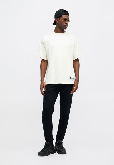Light beige short-sleeve t-shirt with a relaxed fit and a label on the lower hem, paired with black sweatpants and black sneakers.
