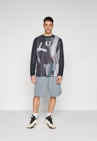 Langærmet sports skjorte i grå camouflage mønster, med sorte ærmer og rundt logo. Grå atletik shorts. Hvide sokker og sorte sneakers fuldender looket.