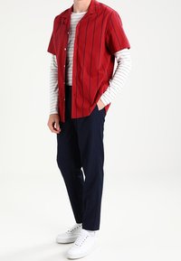 Rode korte mouw knoopshirt met verticale marineblauwe strepen over een wit gestreept langemouwtop. Gecombineerd met marineblauwe broeken en witte sneakers.