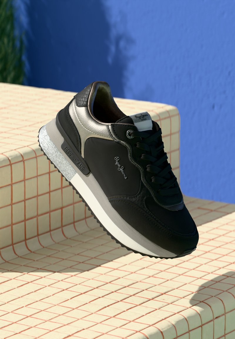 Baskets Pepe Jeans noires et argent avec semelle blanche reposant sur des marches carrelées beige devant un mur bleu.