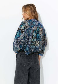 Blouse ample, à motifs dans des tons de bleu avec des designs floraux et géométriques ; associée à un pantalon large taille haute gris foncé.