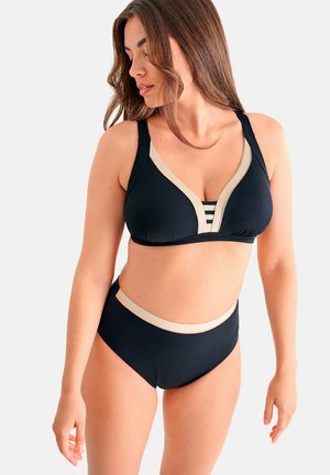 Vrouw met lang haar die een zwarte bikini draagt met beige bies en gestreepte details, staand tegen een effen lichte achtergrond.