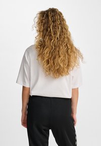 Biały t-shirt z krótkim rękawem wykonany z bawełny, luźny krój, w połączeniu z czarnymi spodniami z białymi paskami po bokach. Widoczne teksturowane kręcone włosy.