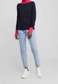 Person som bär en marinblå och klar rosa lager-på-lager långärmad tröja, ljusblå jeans och vita sneakers som står mot en enfärgad grå bakgrund.