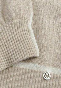 Beige gebreide trui met geribde manchetten, voorzien van een witte horizontale streep en een klein zilveren hartvormig embleem nabij de zoom.