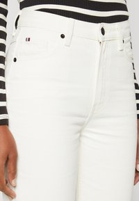 Hvite denimjeans har et klassisk fem-lommers design, svart maskinvare og en liten fargerik logo på frontlommen.