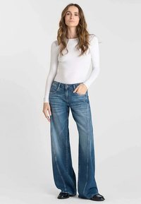 Haut blanc léger à manches longues associé à un jean bleu délavé à jambes larges. Le jean présente des coutures contrastantes et une coupe décontractée. Des chaussures noires complètent le look.