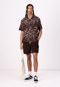 Brun kortermet skjorte med hvit bladmønster, parret med brune cord shorts. Modellen holder en beige tote bag og har på seg svarte sneakers.