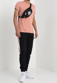 Sac banane noir avec logo blanc, T-shirt rose, joggings noirs avec bandes latérales roses, et baskets blanches ; tissu lisse et coupe décontractée.