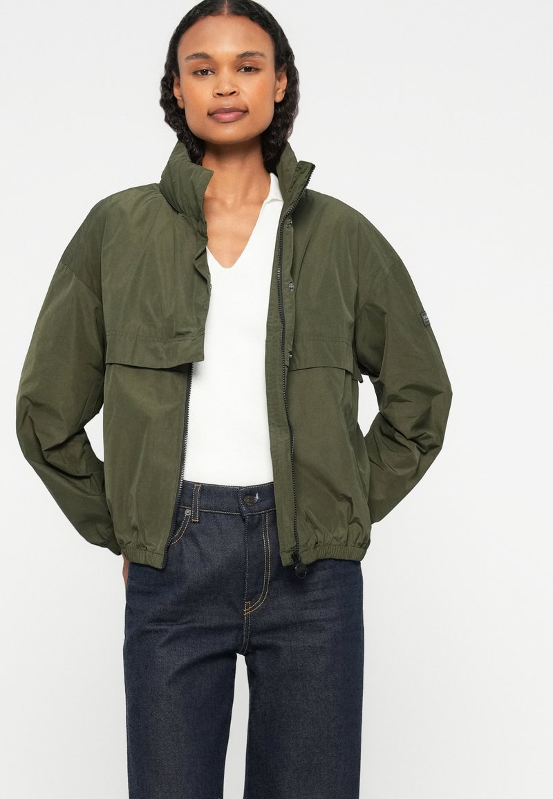 Olivengrüne Zip-Jacke mit hohem Kragen und elastischen Bündchen, über einem weißen V-Ausschnitt-Oberteil getragen, kombiniert mit dunkelblauen Jeans.