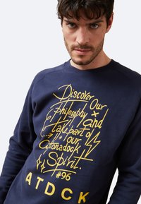 Sudadera azul marino con texto gráfico amarillo y símbolos en la parte frontal. Cuenta con un cuello redondo y puños acanalados. Tejido suave y texturizado.