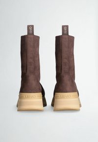 Stivaletti alla caviglia in suede marrone con gambale alto, suola in gomma beige e dettagli marrone chiaro; presentano un'etichetta di marca per facilitare l'indossamento.