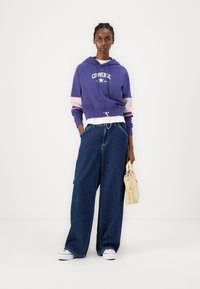 Moletom cropped roxo com mangas listradas em rosa e azul-marinho, apresentando um logotipo branco. Combinado com calças de ganga azuis de perna larga e uma bolsa bege.