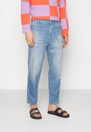 Personne portant un jean bleu clair, des sandales noires à double bride et un pull court à carreaux orange et violet, debout devant un fond uni.