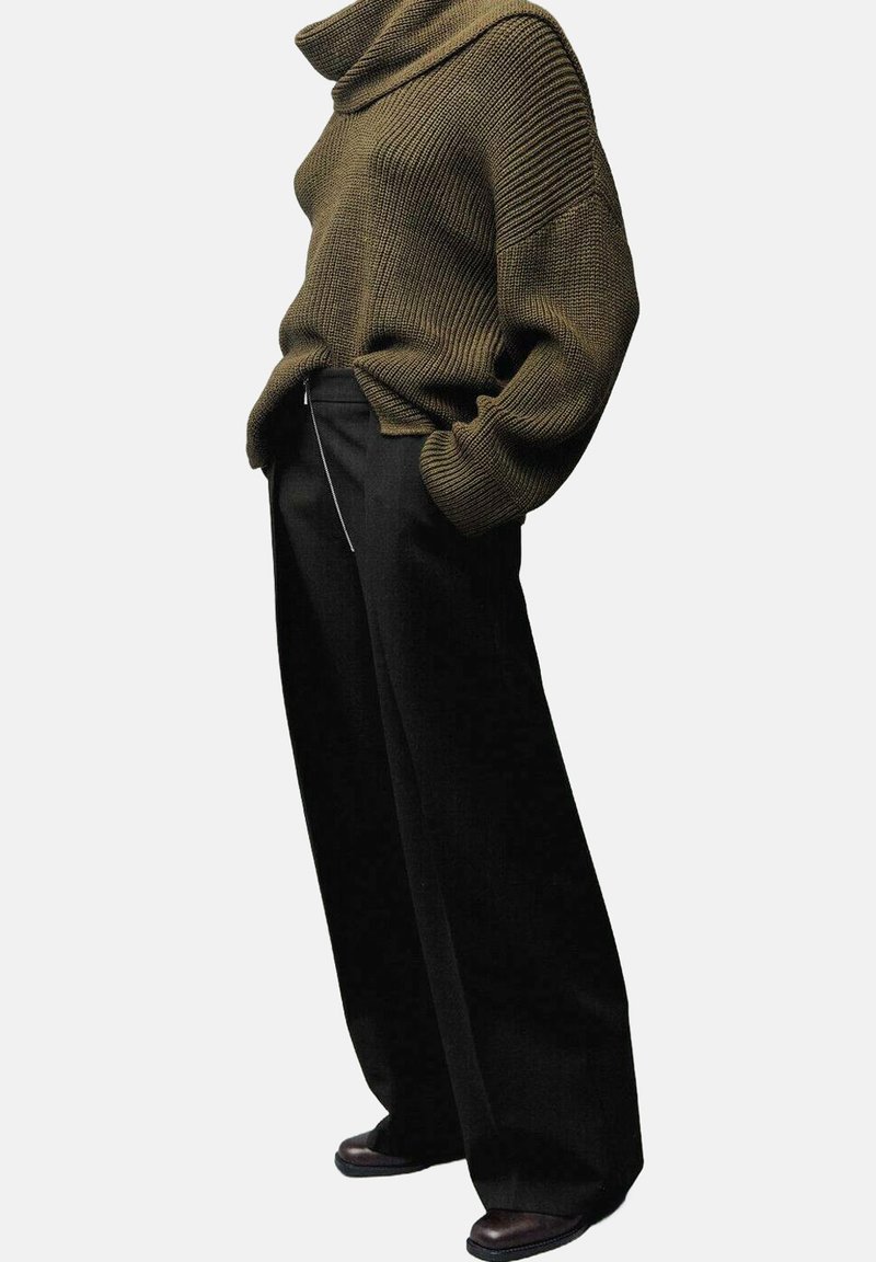 Suéter de punto verde oliva con un alto cuello de cowl, combinado con pantalones de pierna ancha negros y zapatos oscuros. Presenta un detalle de cremallera visible.