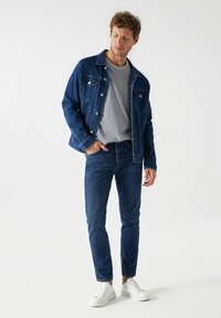 Chaqueta de mezclilla azul sobre una camiseta de rayas gris y blanca, combinada con jeans azul oscuro y zapatillas blancas. El hombre está de pie de manera casual, con la mano en el bolsillo.