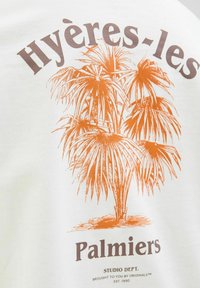 Valge puuvillane t-särk, millel on oranž palmi graafika, peal kirjaga "Hyères-les Palmier" ja all "STUDIO DEPT." pruuni värviga.