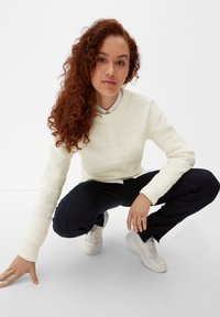 Witte textuurtrui met een ronde hals, gecombineerd met donkerblauwe broek en witte sneakers. Persoon zit in een hurkhouding met los, krullend haar.