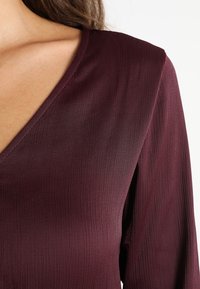 Primer plano de una mujer que lleva una blusa de punto burdeos con escote en V y mangas largas, dejando ver el hombro y la parte superior del pecho.