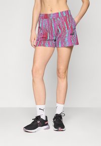 Calças de desporto PUMA com um padrão vibrante em rosa e ciano, tecido leve, cintura elástica e bolsos laterais, combinadas com sapatilhas pretas.