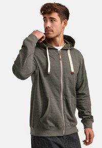 Sudadera con cremallera en verde oliva, con cordones en color crema, tela texturizada, bolsillos tipo canguro, puños acanalados y cierre de cremallera con acentos metálicos.