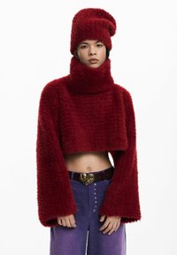 Pulover roșu scurt din material plușat, cu guler înalt și mâneci largi, asortat cu o pălărie similară. Poartă pantaloni mov cu talie înaltă și o curea decorativă.