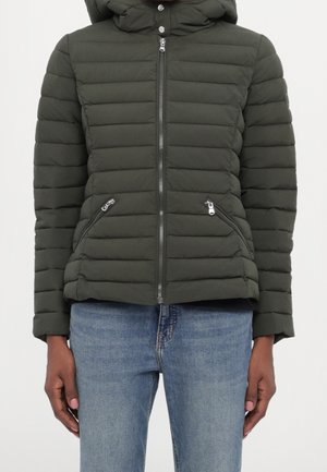 Kobieta ubrana w ciemnozieloną pikowaną kurtkę zapinaną na zamek z ukośnymi kieszeniami na zamek i kapturem, w połączeniu z niebieskimi dżinsami.