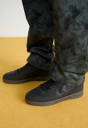 Chaussures de sport noires avec un dessus en cuir lisse et des accents texturés, associées à un pantalon à motif camouflage sombre, sur fond jaune.