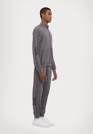 Completo sportivo grigio con giacca con zip e tre righe sulle maniche, abbinato a pantaloni coordinati, completato da sneakers bianche.