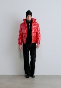 Piros párnázott dzseki fényes felülettel, kapucnival és elől cipzárral, fekete garbó felett viselve, fekete nadrággal és cipővel párosítva.