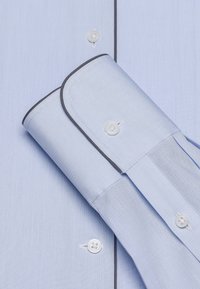 Lichtblauw overhemd van stof met een gladde textuur, knoopsluitingen, contrasterende donkere piping op de manchetten en een opgevouwen mouwdetail.