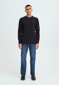 Man die tegen een effen achtergrond staat, gekleed in een zwarte langlopende trui, blauwe jeans en zwarte schoenen, met een ontspannen houding naar voren gericht.