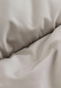 Blødt, krøllet stof i lyse beige nuancer, der viser en glat tekstur med subtil glans og fine folder, som fremhæver dets lette materiale.