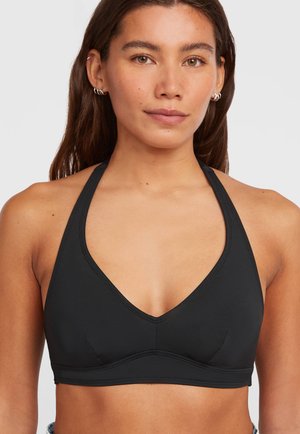 Donna con capelli castani lunghi che indossa un reggiseno sportivo nero a collo alto e piccoli orecchini a cerchio argento su uno sfondo chiaro uniforme.