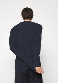 Marinblå ribbad tröja med långa ärmar och rund hals, med en texturerad stickad design och distinkt sömdetaljering längs axlarna.