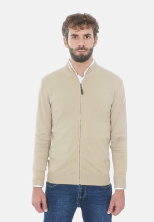 Uomo con barba che indossa un maglione beige con zip sopra una camicia bianca e jeans blu, in piedi di fronte a uno sfondo bianco.