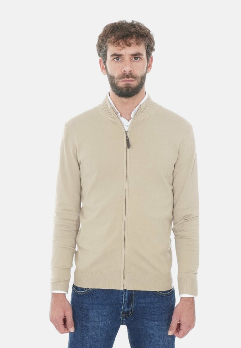 Uomo con barba che indossa un maglione beige con zip sopra una camicia bianca e jeans blu, in piedi di fronte a uno sfondo bianco.