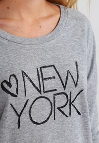Persona che indossa una maglietta grigia con testo nero "New York" e un simbolo a forma di cuore sul petto, mostrando parte del collo e della zona del colletto.