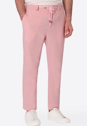 Uomo che indossa pantaloni chino rosa chiaro con cintura e coulisse, sneakers bianche e camicia bianca parzialmente visibile.