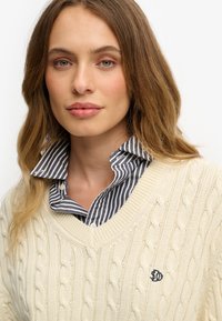 Suéter de punto trenzado color crema con escote en V y logotipo bordado, superpuesto a una camisa de cuello a rayas en azul marino y blanco.