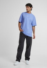 Light blue cotton t-shirt with a relaxed fit and short sleeves, paired with dark gray pants and white sneakers. Simple logo detail on chest.