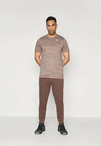 Braunes, gemustertes, kurzärmliges Sportoberteil mit rundem Halsausschnitt, kombiniert mit braunen, schmal zulaufenden Jogginghosen und schwarzen Sportschuhen.