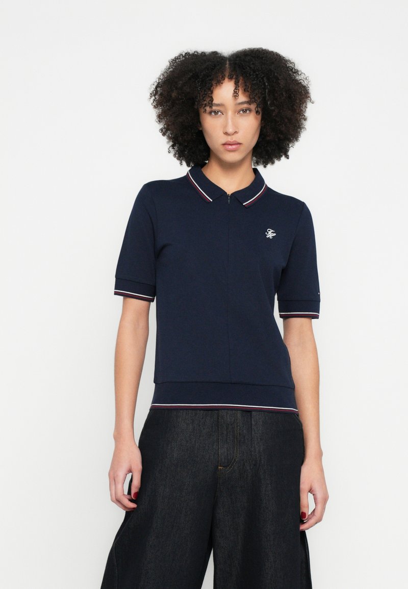 Sötétkék galléros póló cipzáras nyakkivágással és rövid ujjakkal, kontrasztos csíkos díszítéssel és kis hímzett logóval a mellrészen.