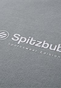 Tissu gris avec texte blanc en relief indiquant "Spitzbub Sportswear Edition" et un logo circulaire sur le côté gauche.