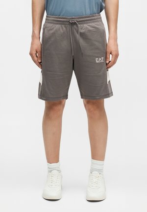 Férfi, aki szürke zsinóros rövidnadrágot visel EA7 Emporio Armani logóval, fehér zoknit és fehér tornacipőt egyszerű háttér előtt.