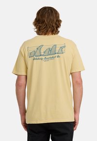 Mężczyzna z włosami do ramion nosi jasnoniebieską koszulkę z nadrukiem rysunkowych finów deski surfingowej, w zestawie zdjęć policyjnych z napisem "Billabong Boardshort Co.".