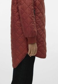 Manteau matelassé rouge avec une poche zippée sur le côté, des poignets côtelés et un ourlet asymétrique. Texture lisse et matériau léger visible contre des jeans noirs.
