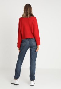 Czerwony, prążkowany sweter z szerokimi rękawami w połączeniu z niebieskimi dżinsami, charakteryzującymi się luźnym krojem, wyraźnymi tylnymi kieszeniami oraz białymi sneakersami.